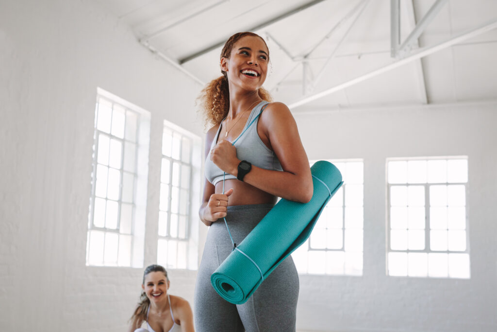 Girl standing in center of frame, holding a yoga mat, looking happy