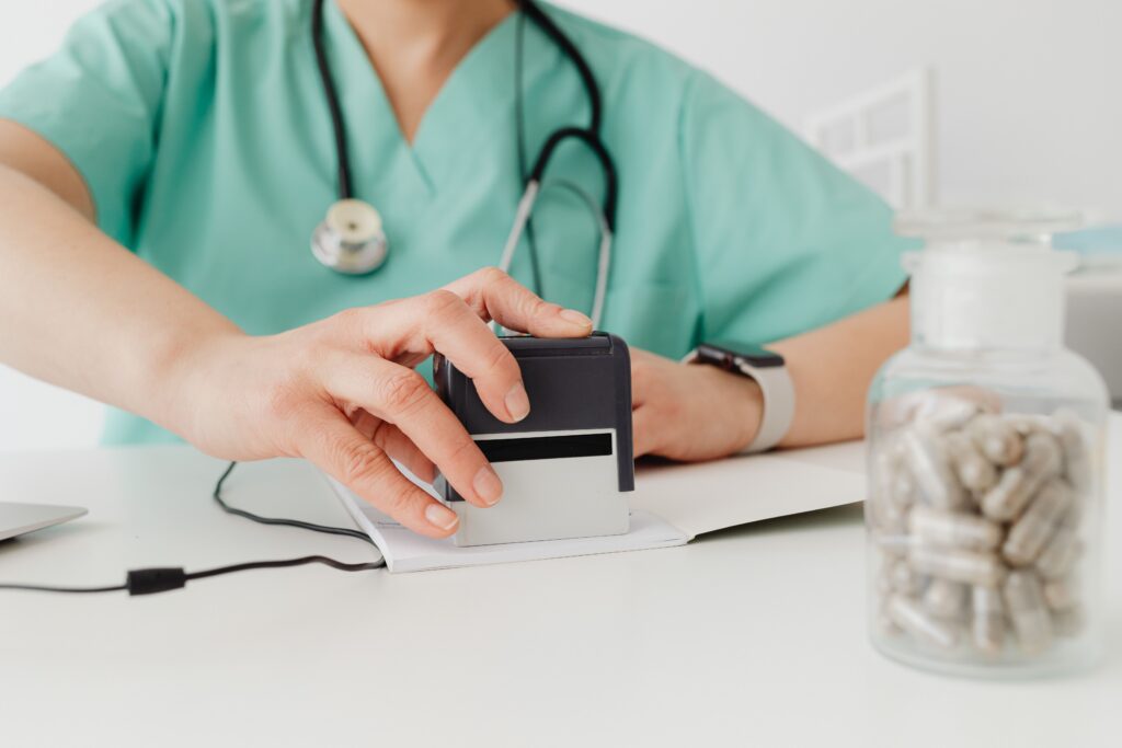 A healthcare worker wearing green scrubs uses a rubber stamp on a piece of paper.