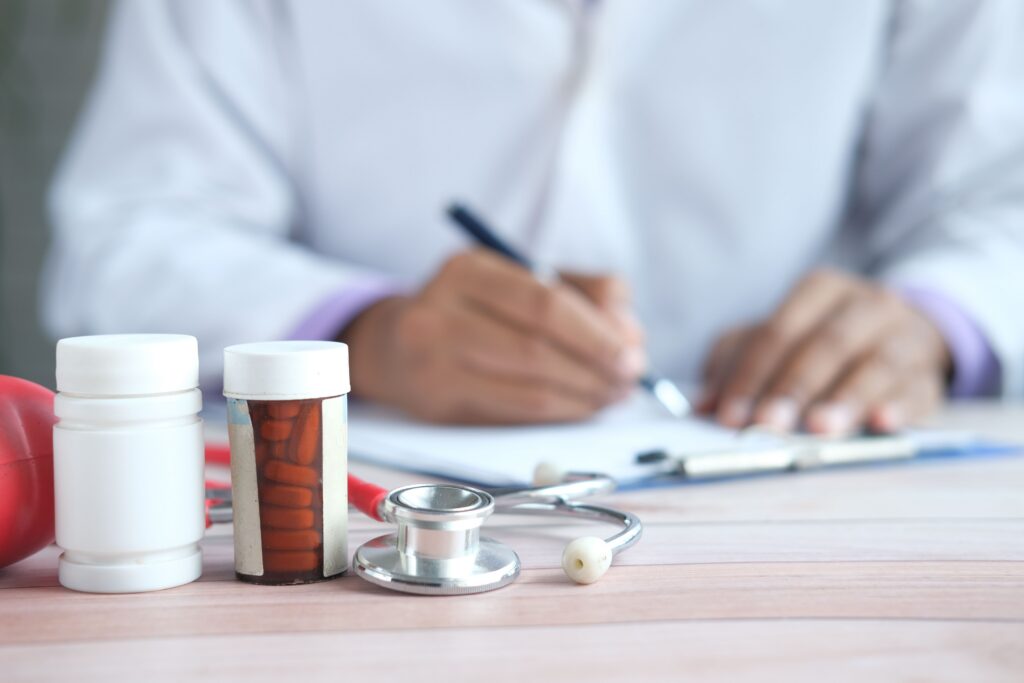 A doctor in a white coat writes on paperwork. Medication tablets and a stethoscope are in the foreground.